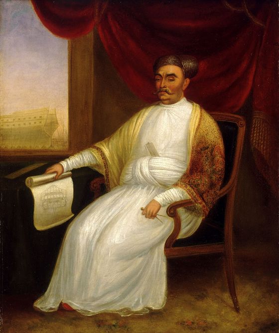 Jamsetjee Bomanjee Wadia (1756-1821), (The shawl, often a gift of the East India Company, may be an addition by the artist.) A famed ship builder, Jamsetjee holds a drawing of the 74 gun ship 'Minden', Launched at Bombay in 1810, the half-built ship can be seen through the window. The Minden was the first man-of-war to be built for the Royal Navy outside of England; in an oil painting attributed to J. Dorman.
